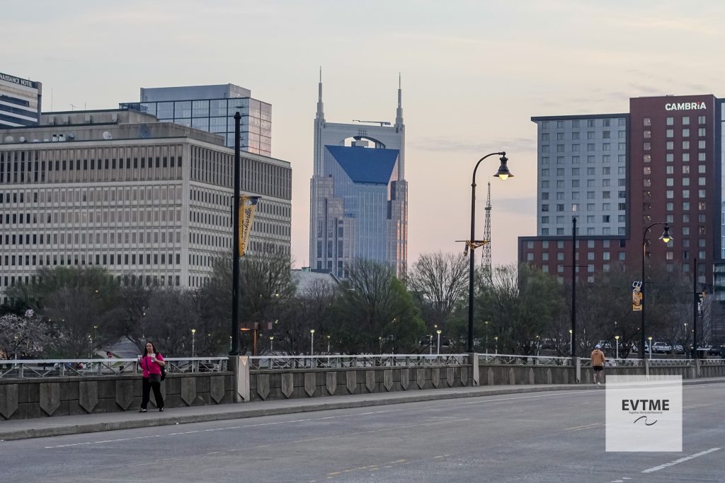 Urban city skyline along a river at dusk, with modern high-rises and a pedestrian promenade in the foreground. A woman in pink walks on the left; a lamppost stands near center. A watermark reads EVTME in the bottom-right corner.