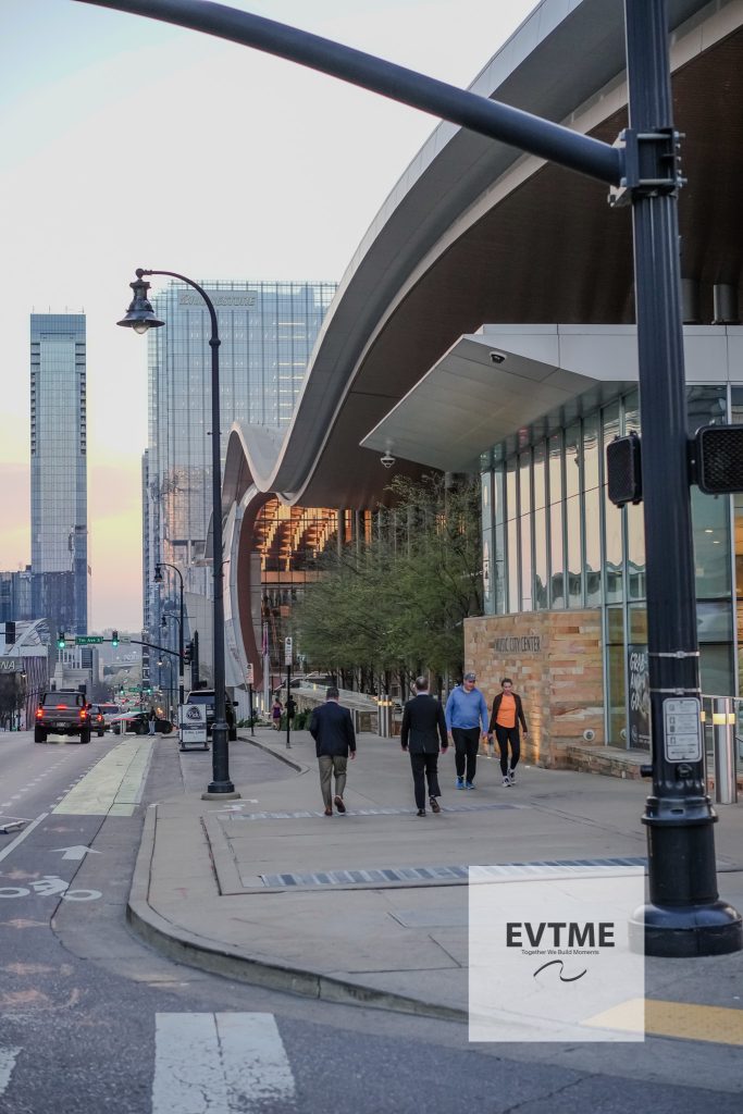 City street with a large curved roof structure and modern glass buildings; pedestrians on the sidewalk and streetlights along the road.