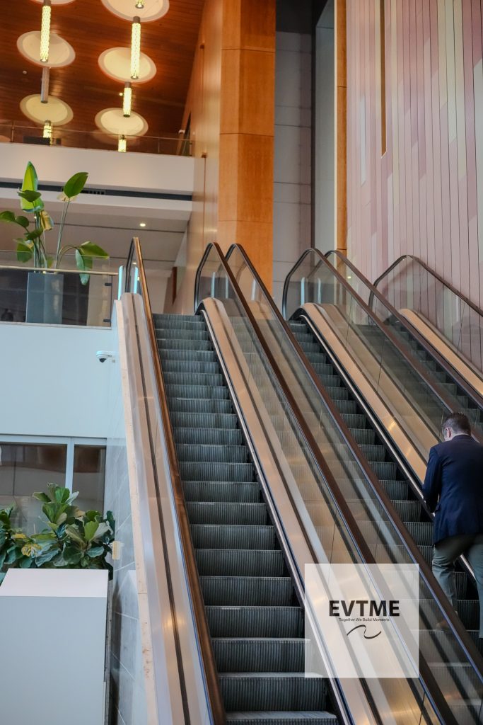 Interior atrium with a pair of escalators, glass railing, and pink panel wall; a person in a suit stands nearby.
