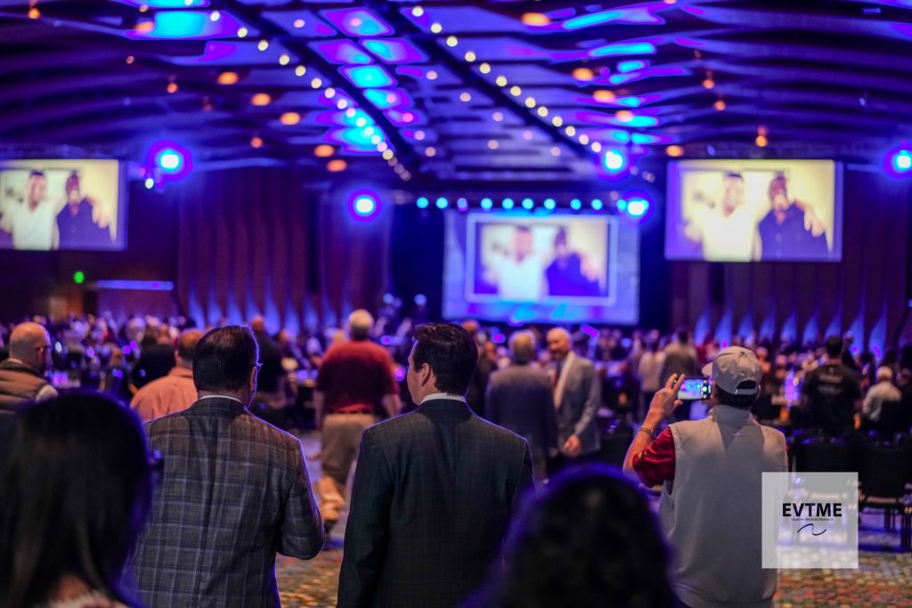 A crowded conference or church hall facing a stage with large screens and blue stage lighting.