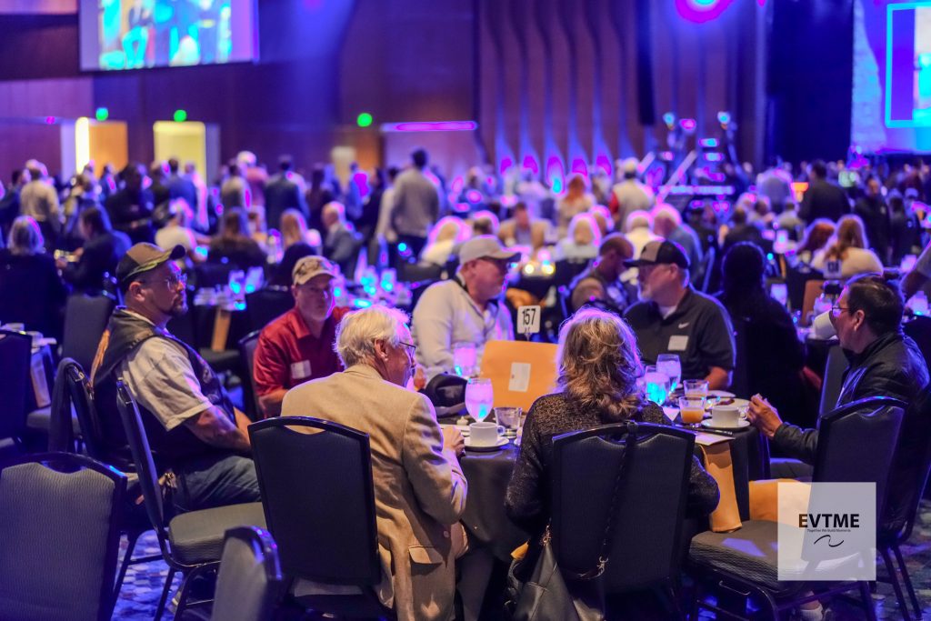 People seated at round banquet tables in a large, blue-lit conference hall; foreground diners chat as drinks and plates adorn the tables; stage area in back with screens visible.