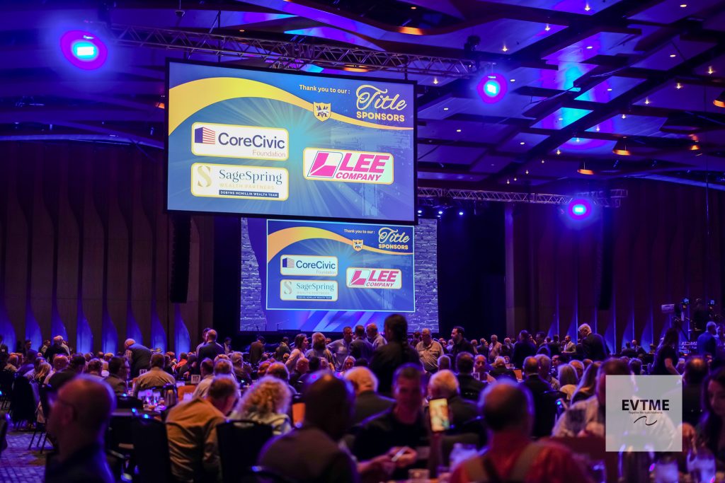 Conference hall awards event with a large screen of sponsor logos (CoreCivic, SageSpring, LEE Company) and purple stage lighting, audience at tables below.