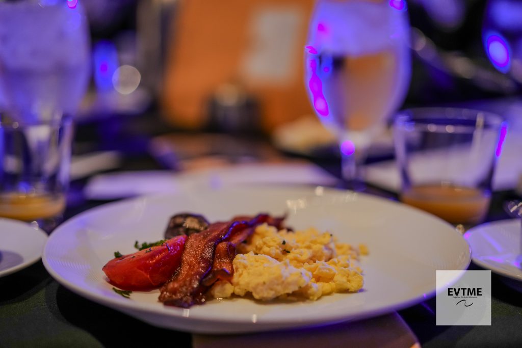 Plate of scrambled eggs with bacon and peppers on a white dish, under blue‑purple restaurant lighting in the background.