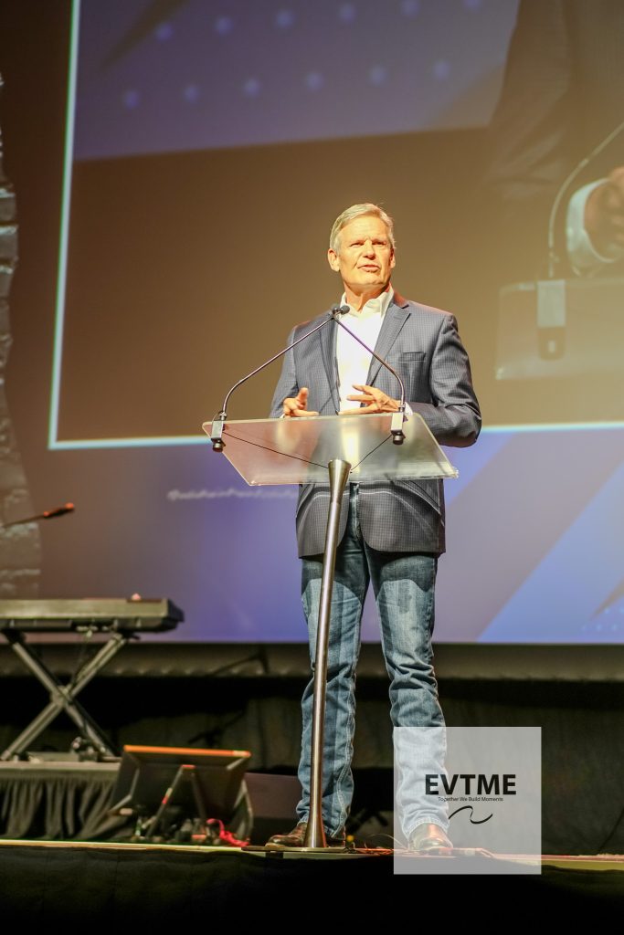 Male speaker in a blue blazer delivers a talk at a clear acrylic podium on a stage with a large screen behind him