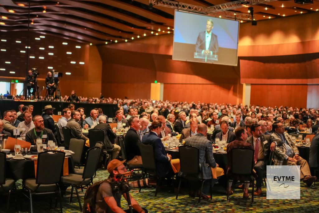 Large conference hall filled with attendees seated at tables, a speaker appears on a large screen above the stage.