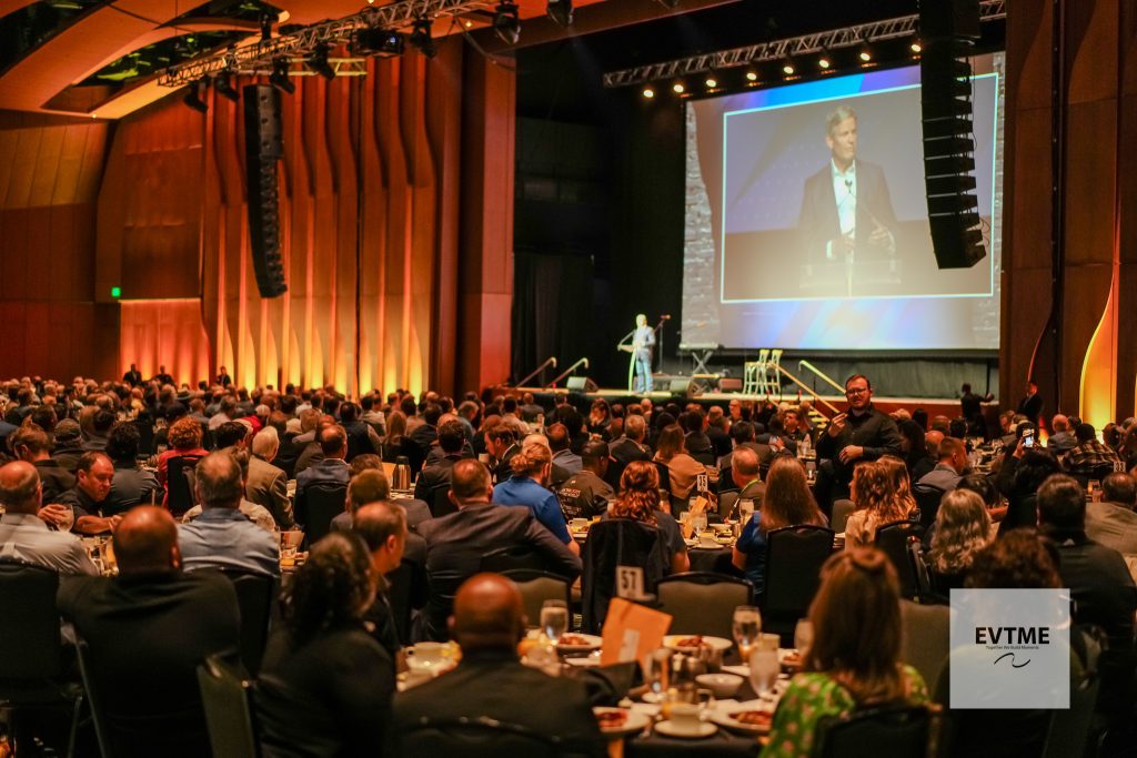 Large audience seated at tables watching a speaker at a podium on stage with a projected slide behind him.