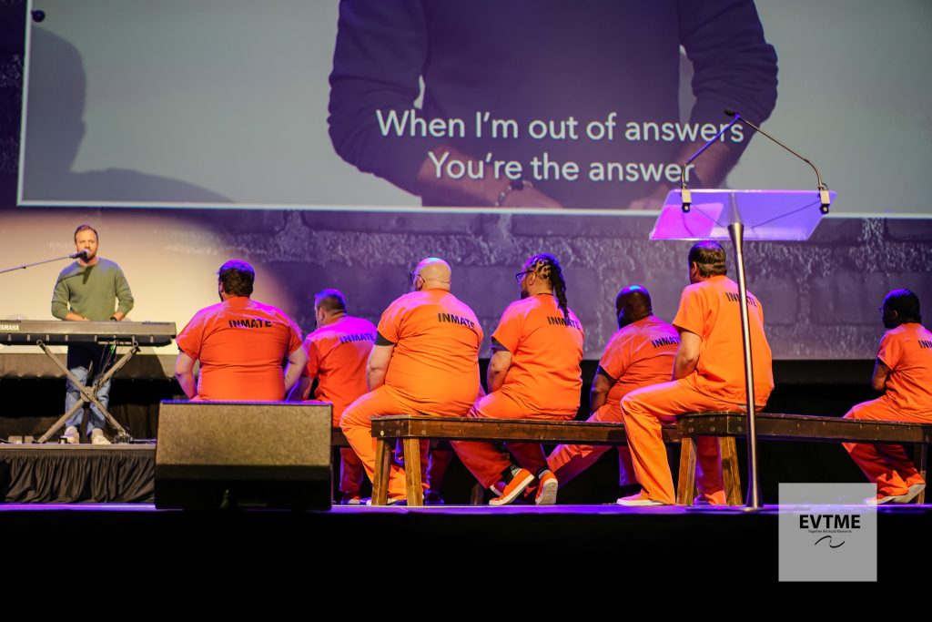 Group of inmates in bright orange uniforms seated on stage facing away, a pianist at left, and a large screen showing the quote “When I’m out of answers You’re the answer.”