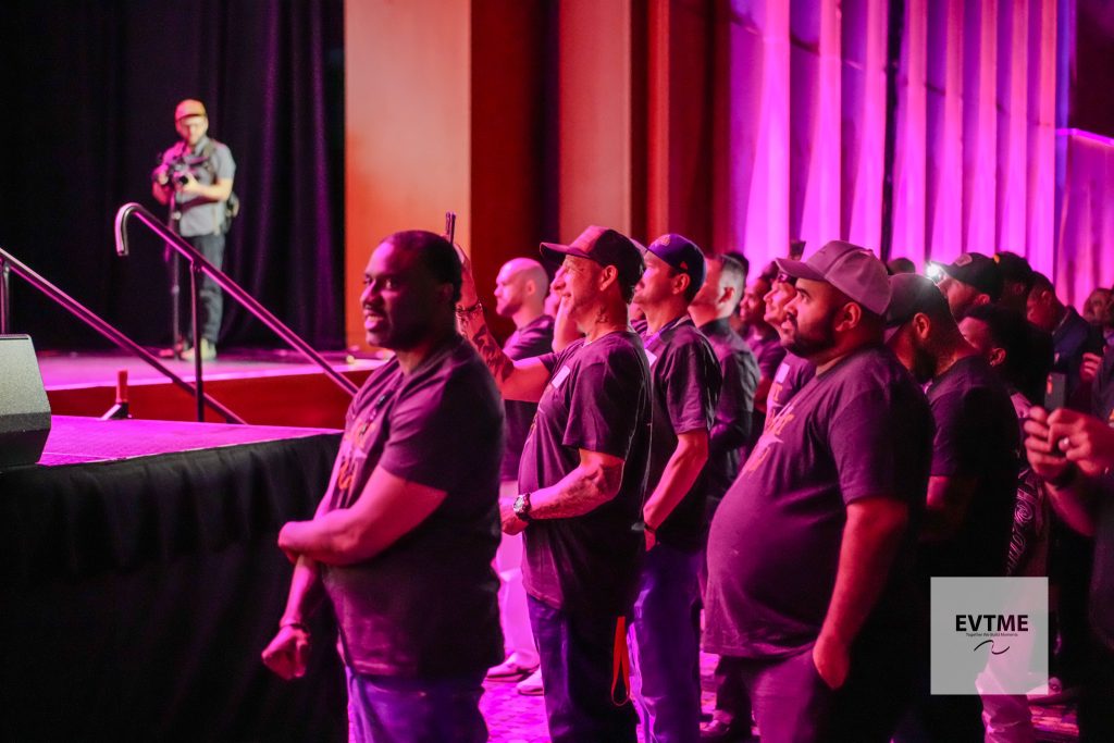 Audience stands along a curb-like barrier facing a stage, bathed in pink lighting, with a photographer on stage left recording the event.