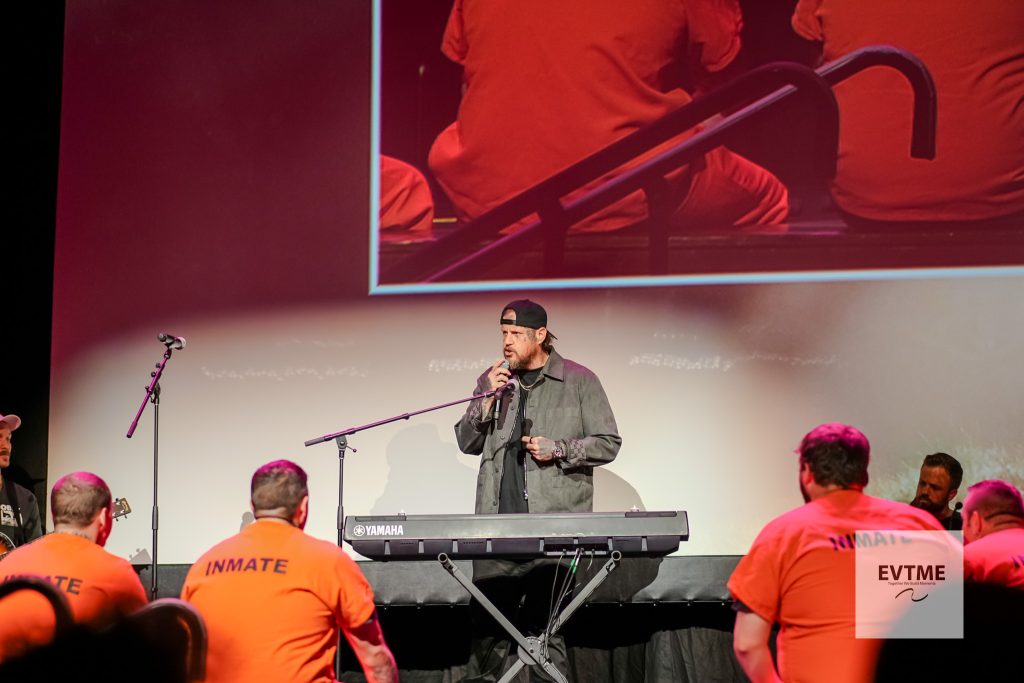 Male performer playing a Yamaha keyboard on stage, microphone in hand, wearing a backward cap, with orange-clad audience in front and a large red screen behind him.