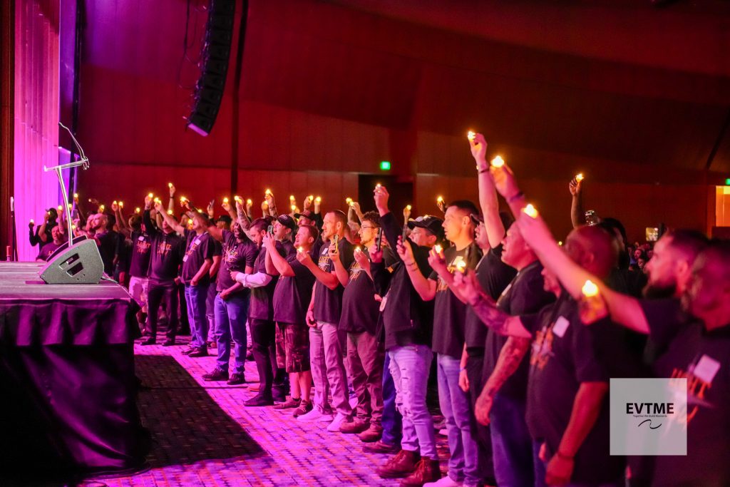 Crowd of people standing in a dim venue, holding lit candles during a performance on stage left, with purple lighting above.