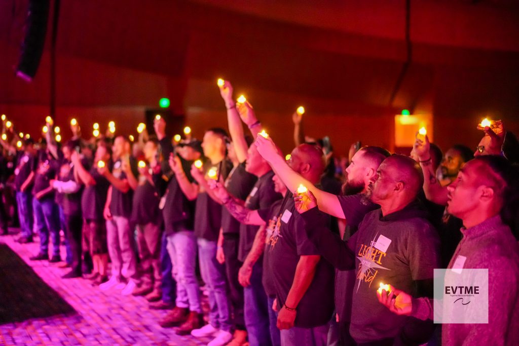 People in a dark venue stand shoulder to shoulder, holding small lit candles aloft in a candlelight vigil/planned event.
