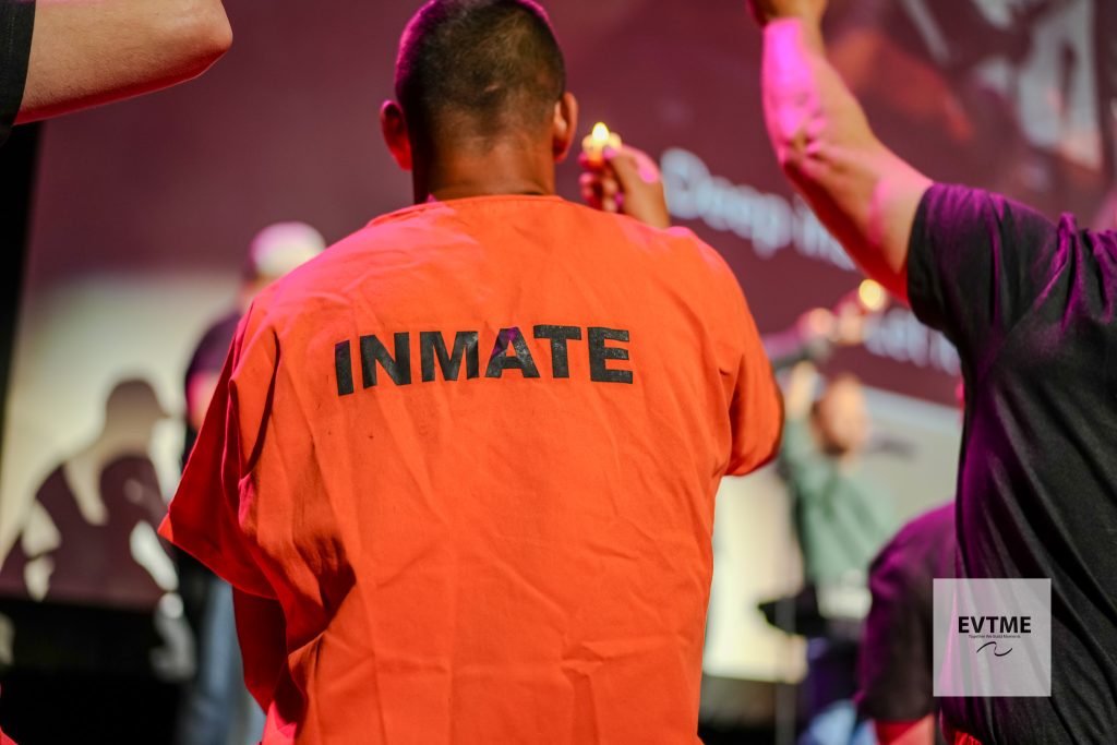 Person seen from behind wearing an orange inmate jumpsuit with 'INMATE' on the back at a stage or event under pink lights.