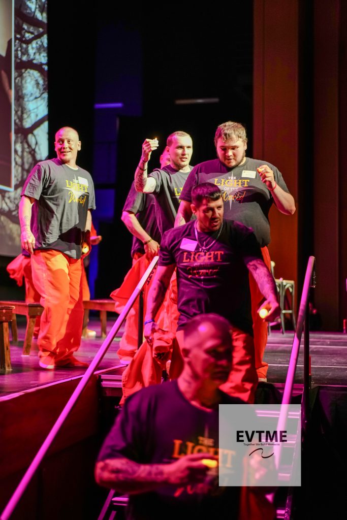Group of men on a stage wearing matching gray shirts that say 'Light of the World' and bright orange pants under pink/purple lighting during a performance.