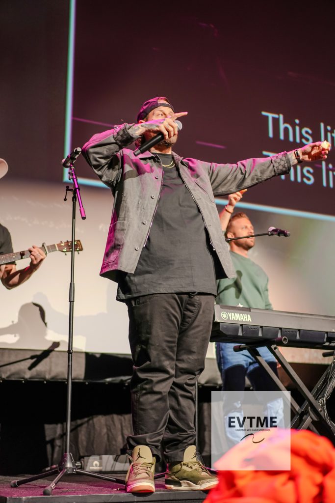 Male vocalist on stage reaching out with microphone, wearing cap and gray jacket, Yamaha keyboard in front and guitarist visible on left.