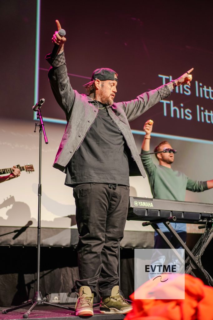 Male performer on stage with microphone, arms raised, wearing a cap and gray jacket beside a Yamaha keyboard.
