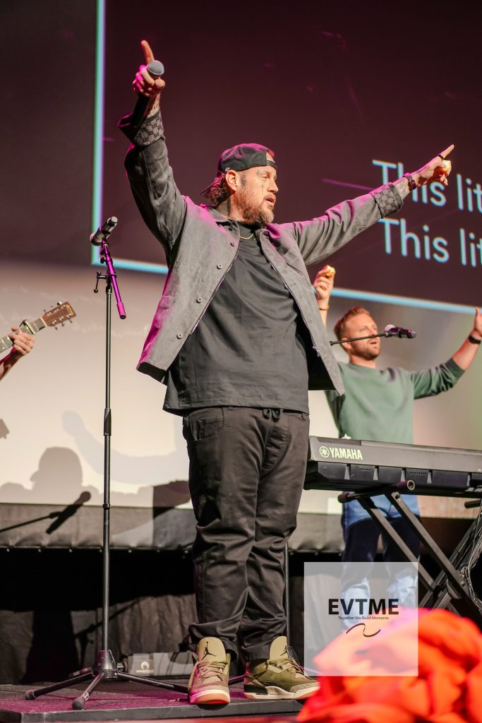 Male performer on stage raises both arms, holding a microphone in his left hand, wearing a gray jacket and cap, near a Yamaha keyboard.