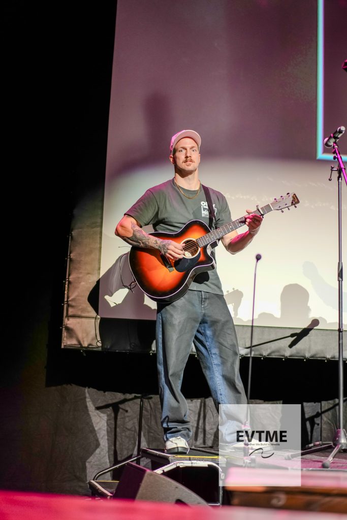 Male guitarist wearing a pink cap and gray T‑shirt, playing a sunburst acoustic guitar on stage.