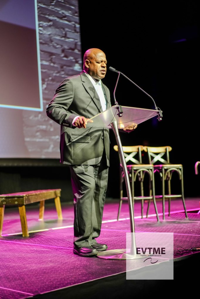 A bald man in a dark suit speaks at a clear podium on a theater stage, with purple stage lighting and a brick backdrop behind him.