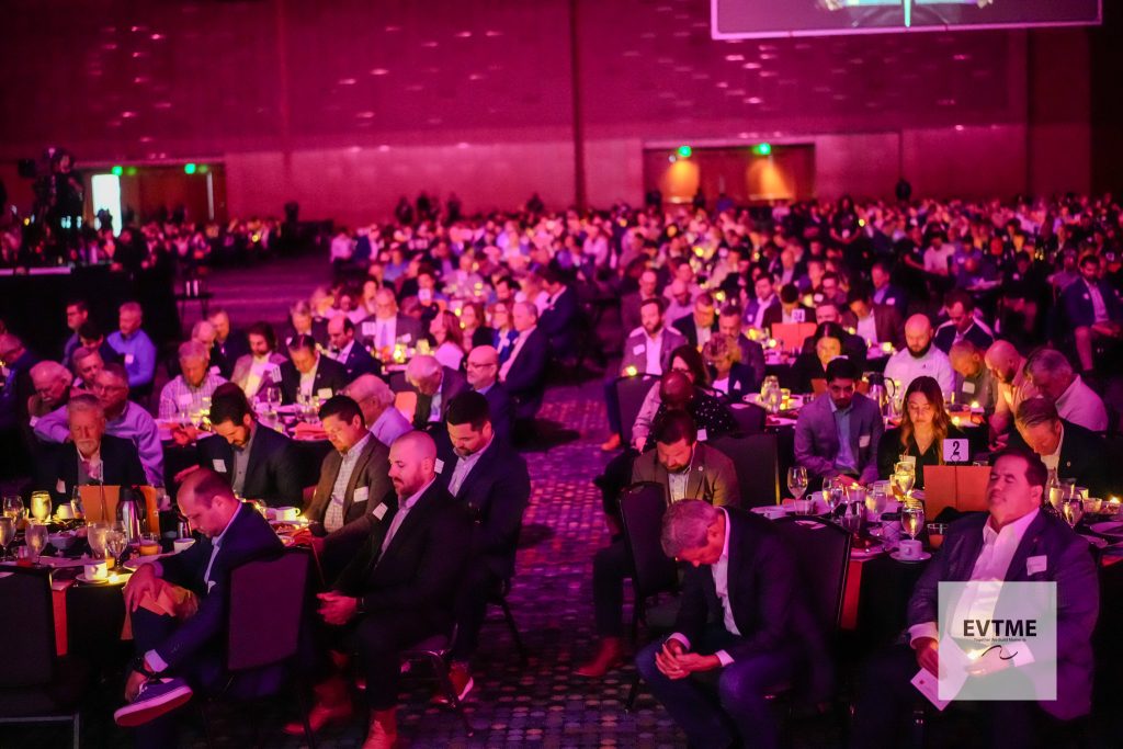 Banquet-style conference with many attendees seated at round tables under pink lighting, likely at a formal event or gala. Watermark bottom right reads EVTIME.