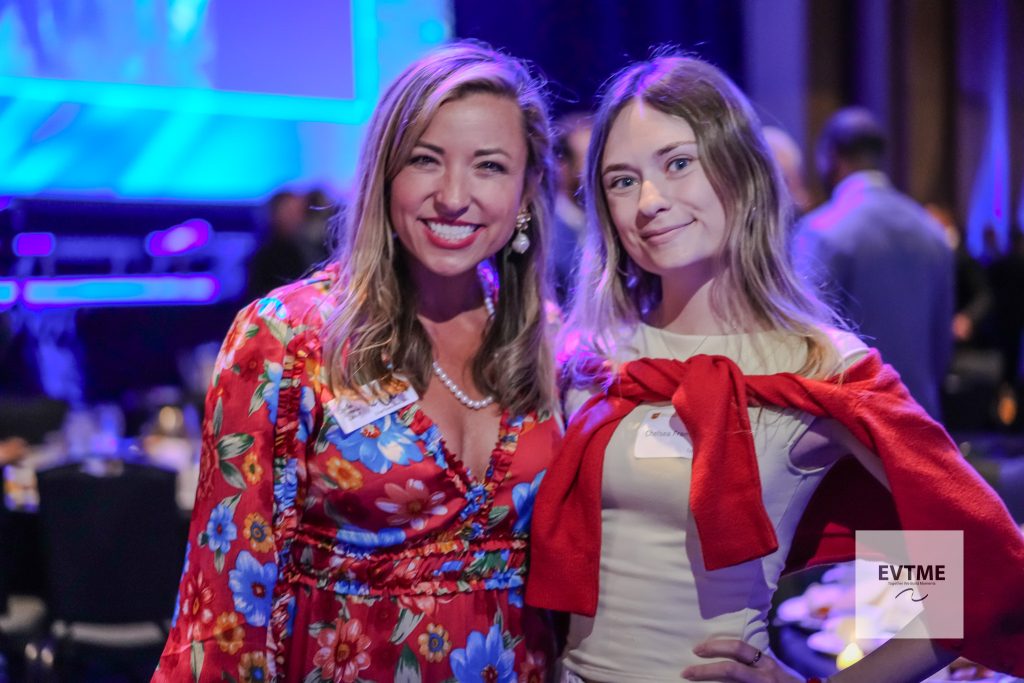 Two women posing and smiling at a party or event, one in a bright floral dress and the other in white with a red sweater draped over shoulders, blue stage lighting in the background.