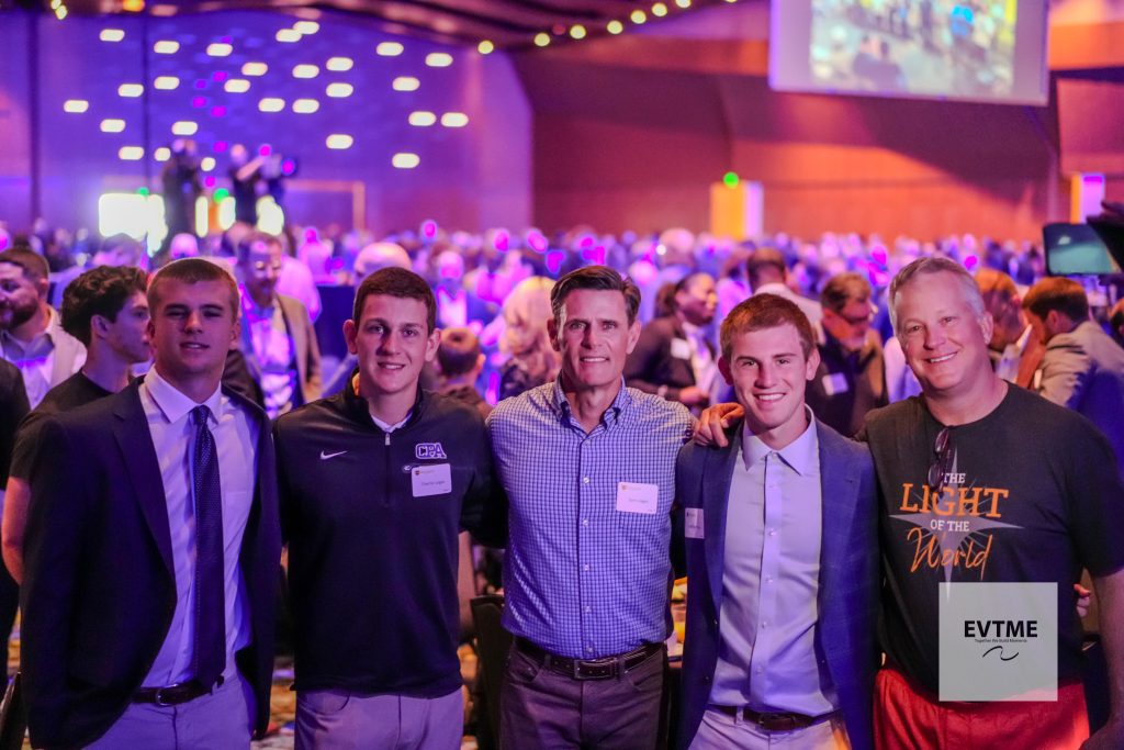 Five men posing for a group photo at a crowded indoor event with colorful stage lights in the background.