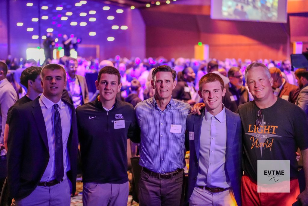 Five men pose for a group photo at a crowded indoor event with purple lighting; they wear business casual clothes and have name badges visible.