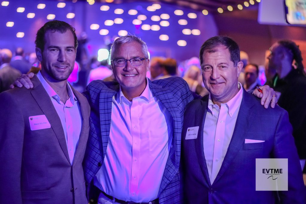 Three men in suits pose together at a dimly lit party, blue-purple lights in the background, name badges visible on their jackets.
