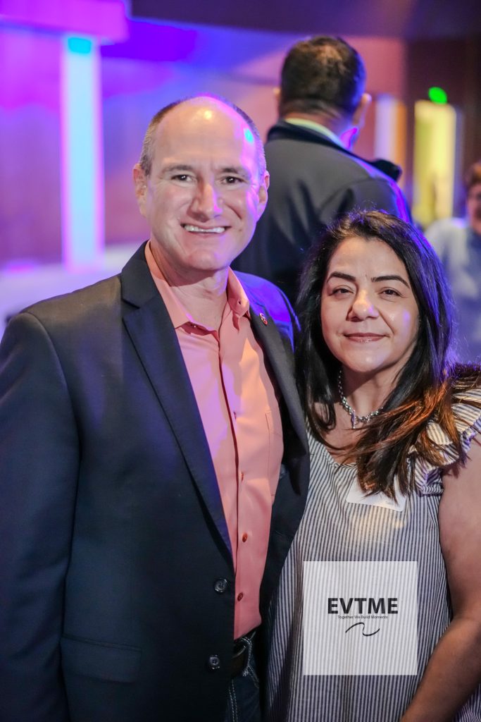 Two smiling adults posing at an event; man in a dark suit and pink shirt, woman in a striped blouse holding an EVTIME brochure.