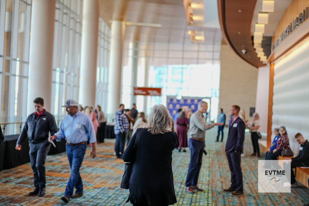 Back of a woman in a dark blazer standing in a crowded conference lobby with attendees chatting in the background.