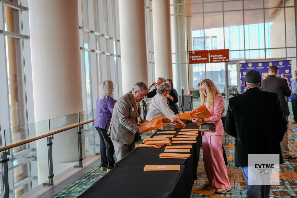 People at a conference registration desk in a bright atrium, distributing orange folders to attendees.