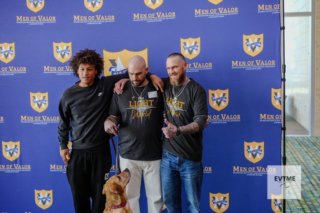 Three men posing together with a golden retriever at a Men of Valor backdrop, arms around each other.