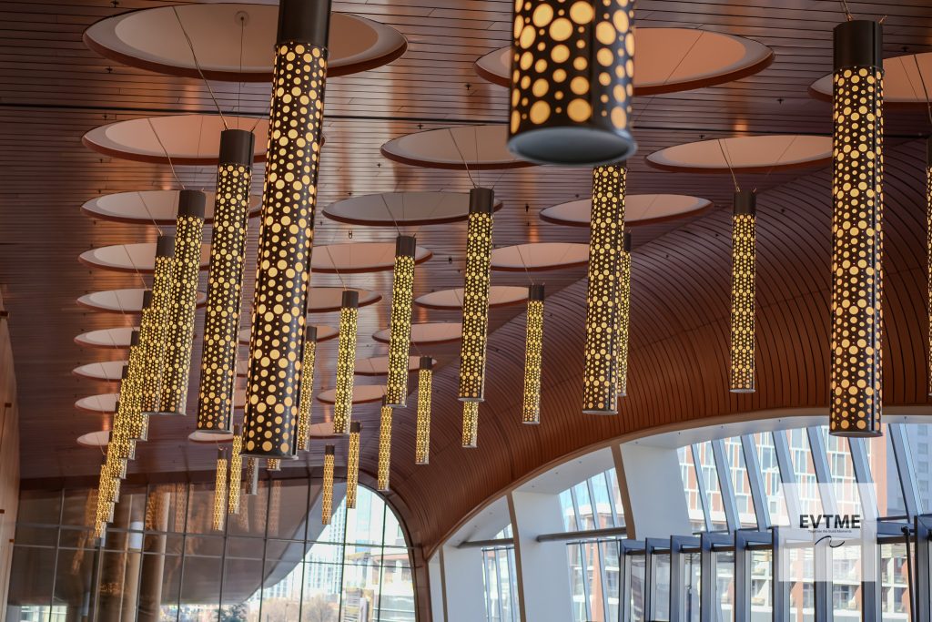 Interior atrium with tall dotted cylindrical pendant lights hanging from a curved wooden ceiling and glass walls.