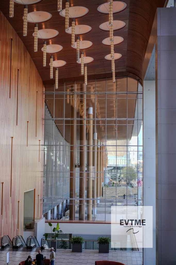Modern lobby/atrium interior with tall hanging light cylinders, curved wooden ceiling, and a glass wall; group of people near escalators.