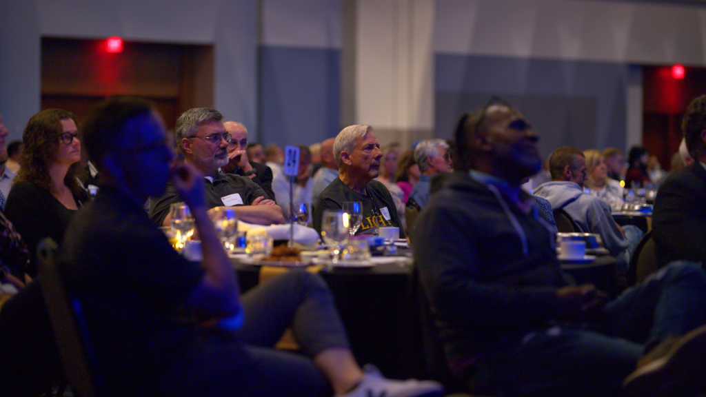 Audience seated at round tables in a dimly lit banquet hall, listening to a presentation at a formal event.