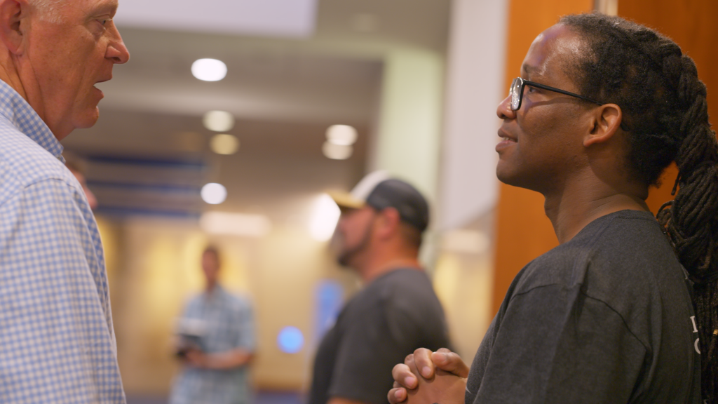Two men face each other in a conversation in a busy indoor space; man on the left wears a blue checkered shirt, man on the right has glasses and dreadlocks.",