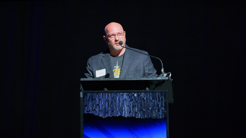Bald man in glasses speaks at a podium on a dark stage, microphone in front of him.