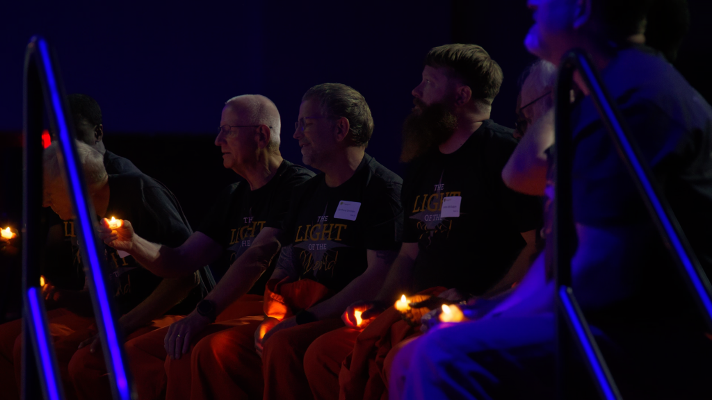 Group of men in matching shirts sit in a dark room, each holding a lit candle as blue neon lights glow nearby.