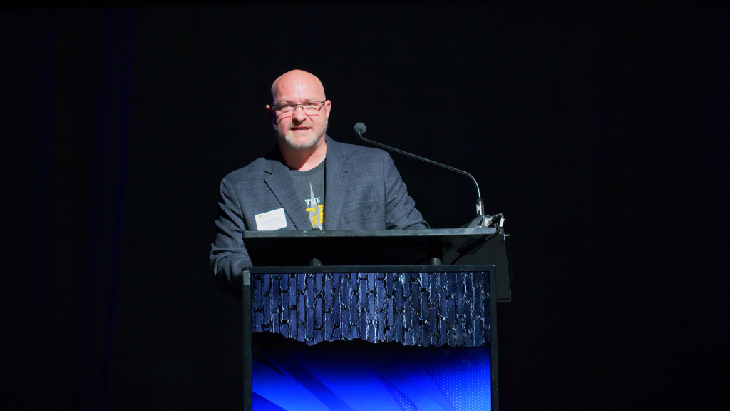 Man with glasses at a podium delivering a talk, microphone on a stand, dark stage behind him.