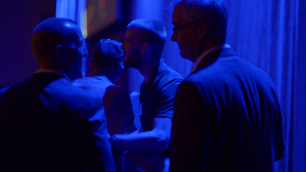 Group of people standing close together in a dark blue-lit room, photographed from behind.