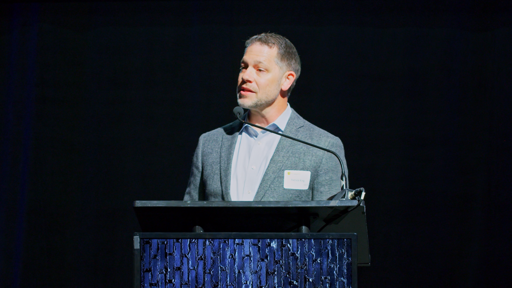 Male presenter in a gray blazer at a lectern on a dark stage, wearing a name badge.